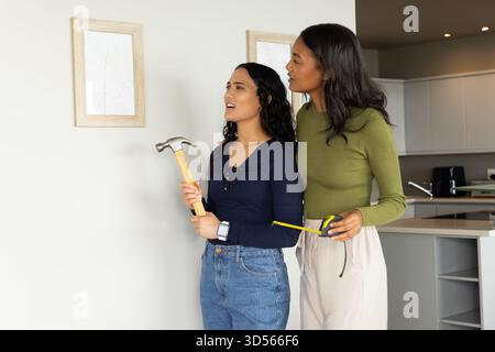 Diverse female friends aligning tape measure on kitchen wall, holding hammer near picture frames Stock Photo