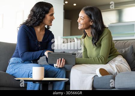 Sharing Diverse female friends browsing tablet on gray sofa in lounge, with coffee mug on table Stock Photo
