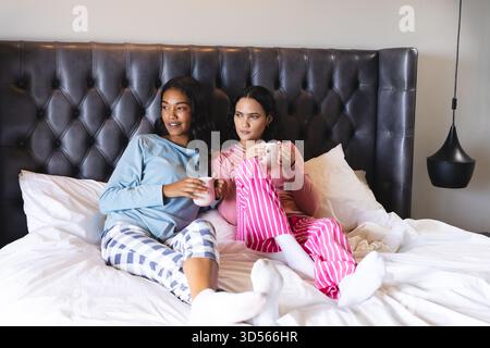 Holding white mugs Diverse female friends sitting on bed in modern bedroom, with leather headboard Stock Photo