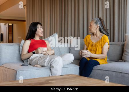 Two Asian women sitting on sofa at home, enjoying coffee and having conversation Stock Photo