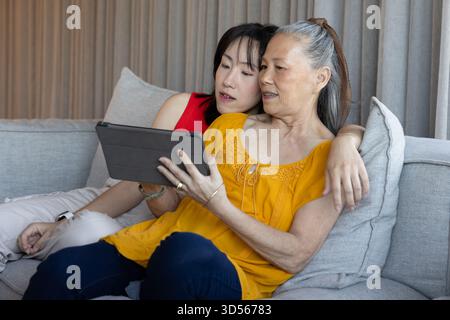 Asian senior mother and daughter using tablet together on couch, sharing joyful moment Stock Photo