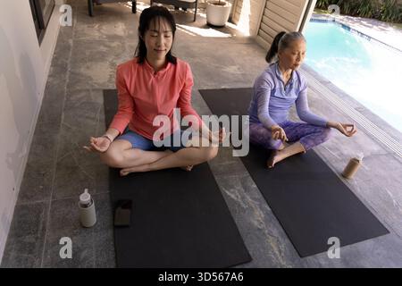 Two Asian women meditating on yoga mats by pool, practicing mindfulness together Stock Photo
