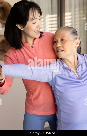 Asian woman helping elderly senior mother with stretching exercises at home, both smiling Stock Photo