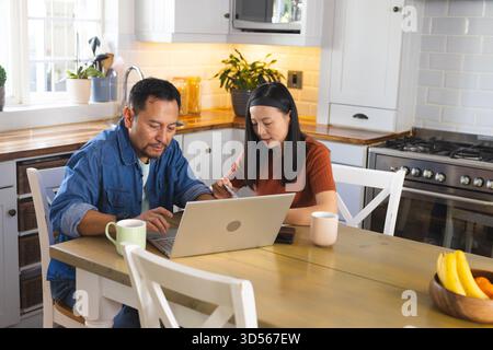 Mature couple using laptop and writing notes at kitchen table, focused on work Stock Photo