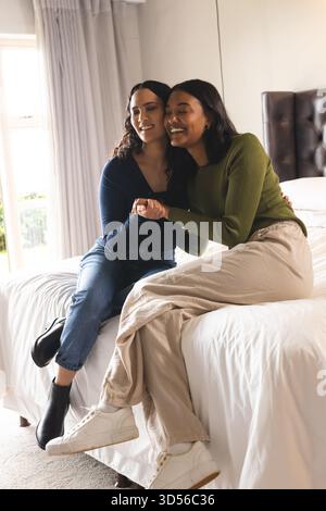 Sitting diverse female couple leaning into each other on bed in lit bedroom, with white linens Stock Photo