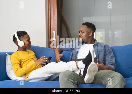 African American father and son talking on sofa, son wearing headphones and smiling Stock Photo