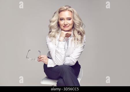 Attractive adult lady with blonde hair, dressed in a stylish casual look, sitting on a chair on a gray isolated background. Stock Photo
