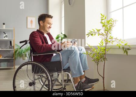 Smiling young man sitting in wheelchair, using digital tablet, making video call or texting online. Stock Photo