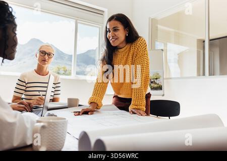 A diverse team of architects, including women of various ages, working together in a well-lit office space, with plans on the table and a sunny window Stock Photo