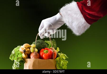 Concept of Christmas grocery shopping with Santa Claus's hand in a suit and gloves holding a paper bag full of food and green isolated background. Stock Photo