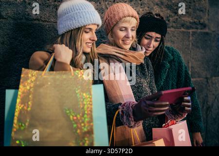 Three happy young friends are looking at a smartphone together while shopping in the city at night during winter. Stock Photo
