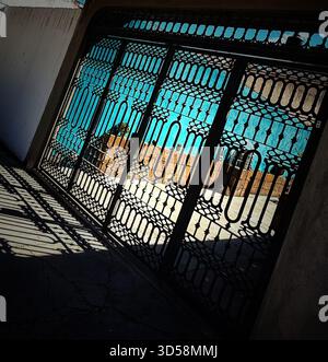 Decorative wrought iron gate casting intricate shadows on concrete in front of a bright blue sky and urban background Stock Photo