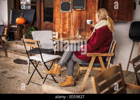 Autumn coffee break at cozy outdoor cafe. Woman in red coat enjoying cup of coffee at street cafe in fall atmosphere, dressed in warm clothes Stock Photo