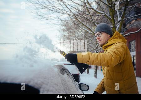 Man in winter clothing clearing snow from car parked in residential city street during cold morning. Scene showing everyday winter routine, weather co Stock Photo