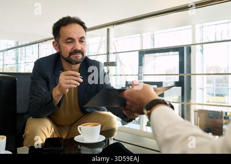 Middle aged Caucasian man sitting at car dealership desk reviewing contract with another , holding pen and clipboard, discussing terms of vehicle purchase over coffee Stock Photo