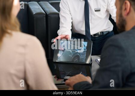 Caucasian young adult man presenting car details on digital tablet to Caucasian young adult woman and Caucasian young adult man at car dealership office table Stock Photo