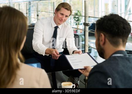 Caucasian young adult man presenting contract to multiethnic young adult couple in modern car dealership, man holding pen and explaining document while couple listening attentively Stock Photo