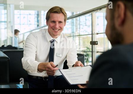 Caucasian young adult man smiling while handing contract to Middle Eastern young adult man in modern car dealership office, both engaged in discussion about vehicle purchase or lease Stock Photo