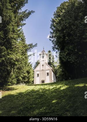 A quaint white chapel with a tower stands quietly among tall green trees on a sunny hillside, surrounded by lush grass and peaceful forest in summer, Stock Photo