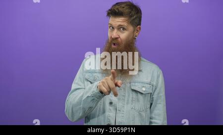 Bearded man in denim jacket with long ginger beard and open mouth expression pressing hand to chest in purple studio; surprise. Stock Photo