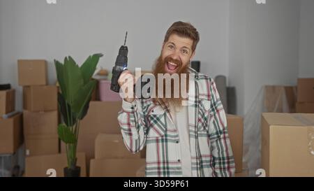 Bearded man holds cordless drill and smiles inside building stacked with cardboard moving boxes in new home; excitement achievement. Stock Photo