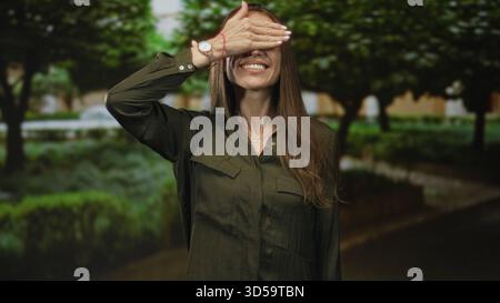 Woman covering eyes with hand and smiling in a forest park with green trees and path, wearing watch and necklace; playful joy. Stock Photo