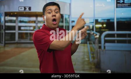 Hispanic man points finger to terminal sign at airport wearing red polo, mouth open in surprised exclamation; surprise curiosity. Stock Photo