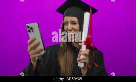 Young hispanic woman wearing mortarboard holds diploma and smartphone for selfie in studio; achievement pride. Stock Photo