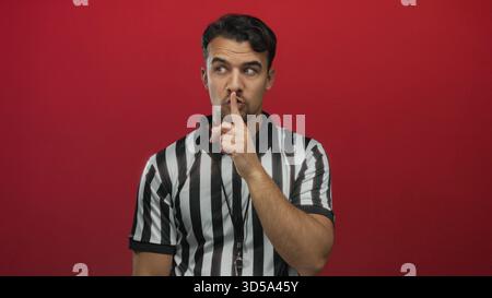 Young hispanic man referee in striped shirt making hush gesture on isolated red background conveying silence and authority. Stock Photo