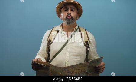 Man reading a folded city map while wearing a backpack and pith helmet in studio with blue backdrop; curiosity adventure. Stock Photo