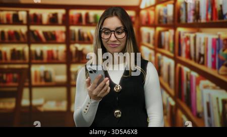 Woman holding smartphone in library with blonde hair and eyeglasses, showcasing a reflective mood under warm lighting among rows of bookshelves indoor Stock Photo