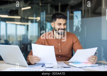 Young businessman analyzing reports and documents at his office desk, confidently reviewing financial data and smiling, showing success and professional growth in a modern workspace Stock Photo