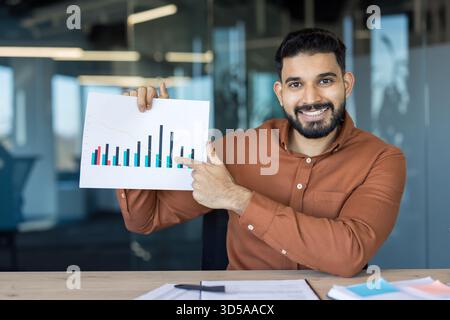 Smiling young indian businessman holding up a graph and pointing, explaining financial statistics during a presentation in a modern office setup, analyzing market trends Stock Photo