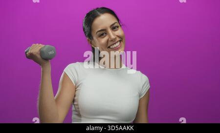 Hispanic woman exercising with a dumbbell against a vibrant pink background, smiling and looking energetic, conveying fitness and positivity in an iso Stock Photo