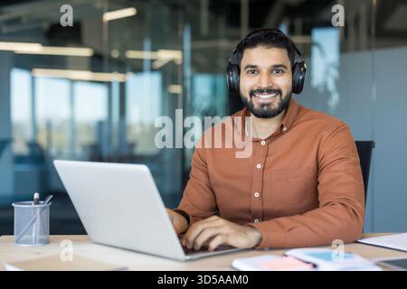 Young indian man in a modern office wearing headphones, smiling at camera while typing on a laptop, engaging in remote work, online learning, or virtual communication Stock Photo