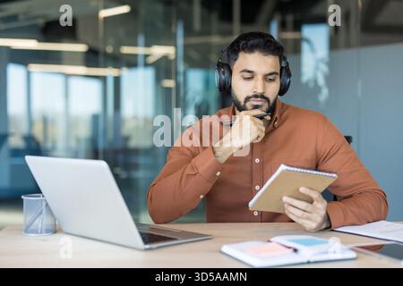 Young man wearing headphones concentrating on online learning while looking at laptop and writing notes in a spiral notebook, sitting at a desk in a modern office Stock Photo