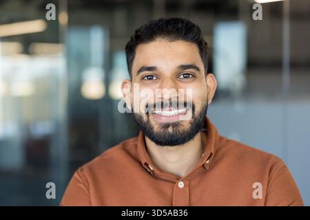 Young indian bearded man smiling confidently, looking towards the camera, standing in a contemporary office environment, representing success, professionalism, and diverse talent Stock Photo