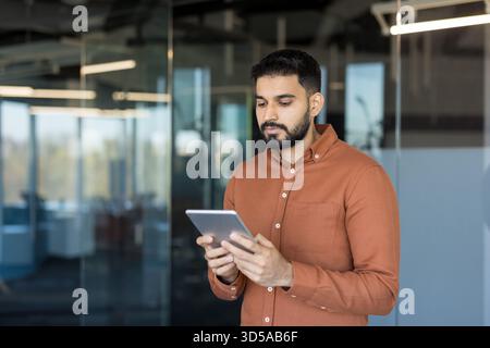 Indian businessman in a brown shirt standing in a modern office, focused on a tablet while analyzing data and using apps for communication and productivity Stock Photo