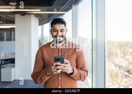 Indian businessman smiling while checking messages on his smartphone, connecting with people, and managing business in a contemporary office building with large windows Stock Photo