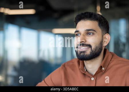 Young adult indian man posing for a half body portrait, confidently smiling and looking away, showcasing professionalism and a positive outlook in a modern office environment Stock Photo