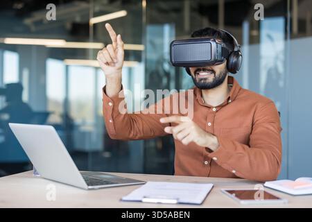 Man wearing a vr headset and headphones, smiling while pointing and interacting with virtual elements, working on future technology and innovation in a modern office Stock Photo