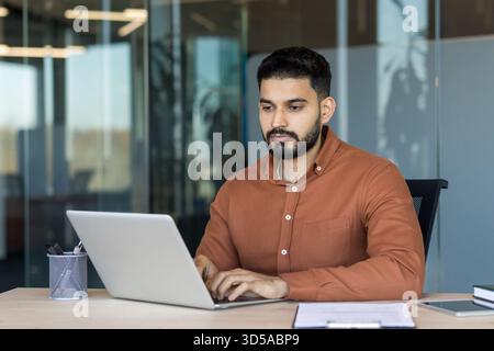 Young indian man staying focused while typing on a laptop at his desk, demonstrating dedication and productivity in a contemporary corporate environment Stock Photo