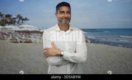 Man with arms crossed and watch on left wrist on a sandy beach with umbrellas and palm trees by the sea; confidence summer leisure. Stock Photo
