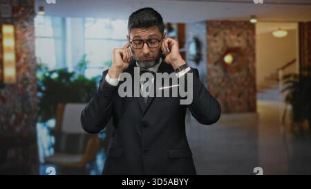 Man in suit adjusting cufflink and buttoning jacket while wearing glasses in building lobby; confidence. Stock Photo