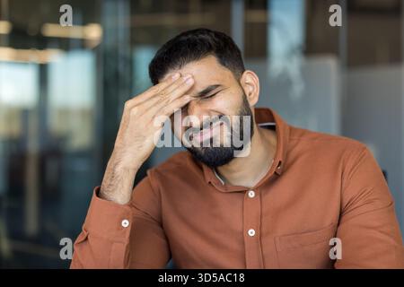 Young man experiencing severe migraine and painful headache, holding hand to his forehead, feeling exhausted and pressured at work in a modern office environment Stock Photo