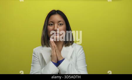 Woman in white jacket looks nervous against a vibrant yellow background, highlighting expressions of uncertainty and hesitance in an isolated studio s Stock Photo
