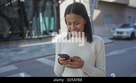 Young hispanic woman using smartphone on sunny street outdoors, smiling confidently as she engages with technology in an urban environment. Stock Photo