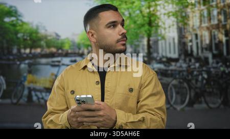 Man in mustard jacket holds smartphone and scrolls screen on street beside canal with parked bicycles and brick buildings; quiet reflection. Stock Photo