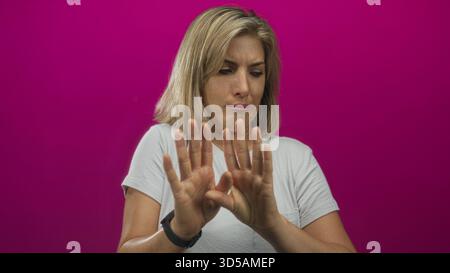 Young blonde woman scrunching nose and raising outstretched hands for stop gesture in pink studio; disgust. Stock Photo