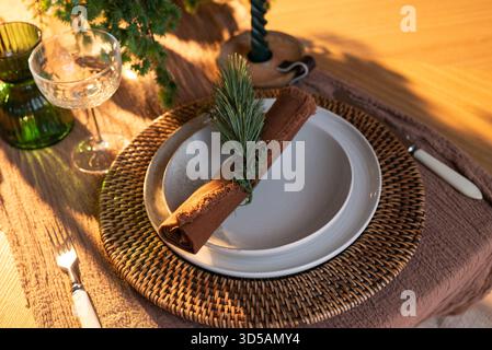Festive table setting with woven charger, ceramic plates, rustic napkin and greenery decoration in warm holiday lighting Stock Photo